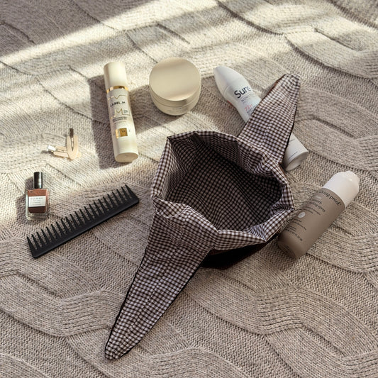 An empty styled flat-lay chocolate brown corduroy make up beauty bag with a dark brown gingham lining with beauty products scattered around. Poetcore aesthetic.
