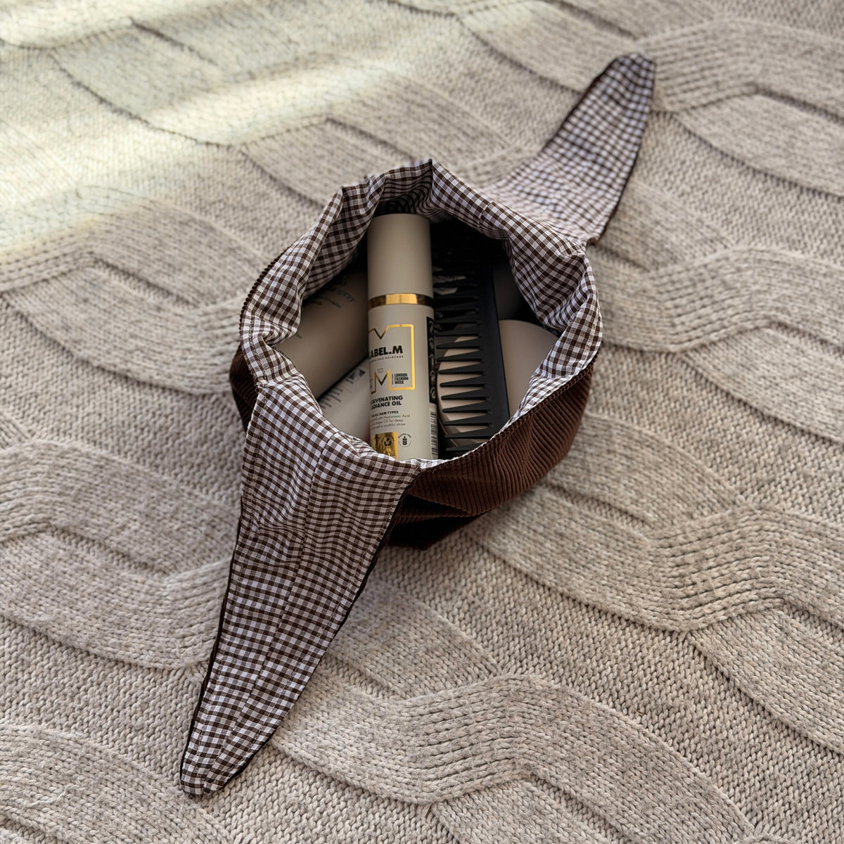A filled styled flat-lay chocolate brown corduroy make up bag with a dark brown gingham lining with beauty products scattered around. Poetcore aesthetic.