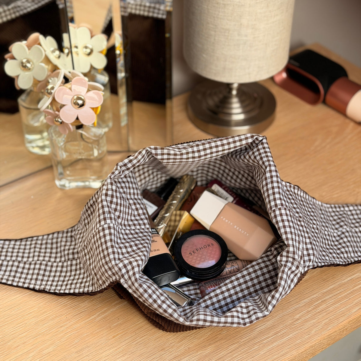 A chocolate brown corduroy and brown gingham makeup bag filled with cosmetics, placed on a wooden desk next to a lamp and mirror. Poetcore aesthetic. 