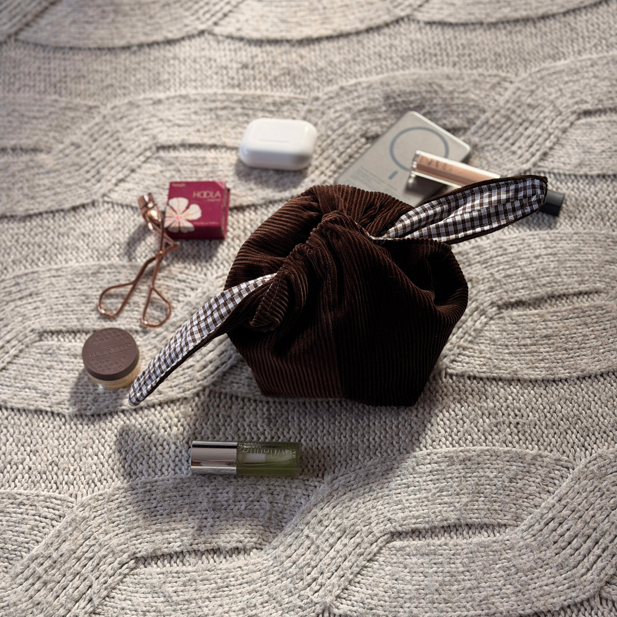 A styled flat-lay chocolate brown corduroy make up beauty organiser bag with a dark brown gingham lining with beauty products scattered around. Poetcore aesthetic. 