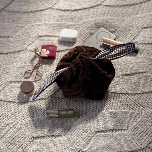 A styled flat-lay chocolate brown corduroy make up beauty organiser bag with a dark brown gingham lining with beauty products scattered around. Poetcore aesthetic. 