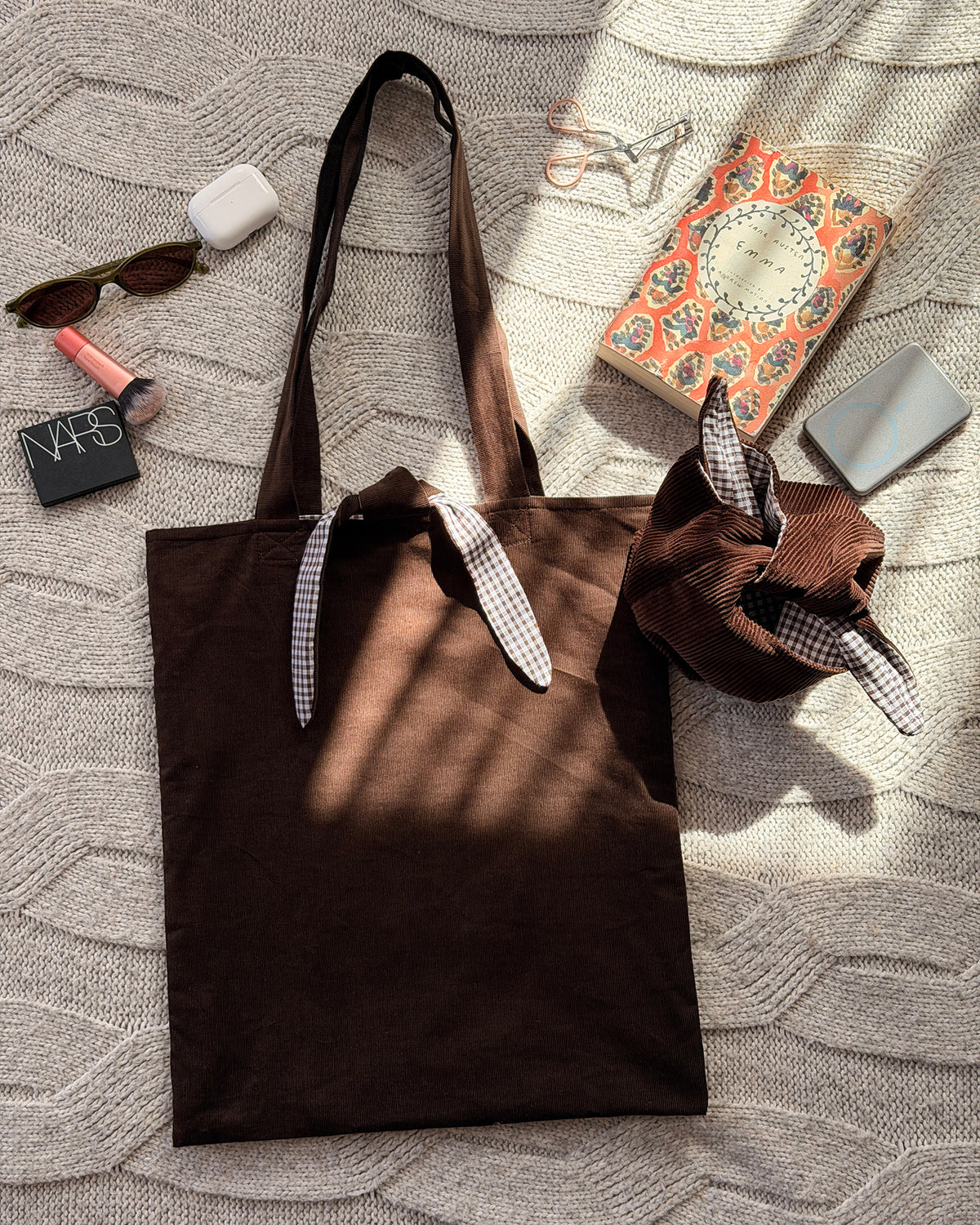 A dark chocolate brown corduroy fabric tote bag with long handles lined with brown gingham and tie and tie detail, with tie up organiser bag to match and beauty products scattered around. Laid flat on a textured cream knit blanket. Poetcore aesthetic.