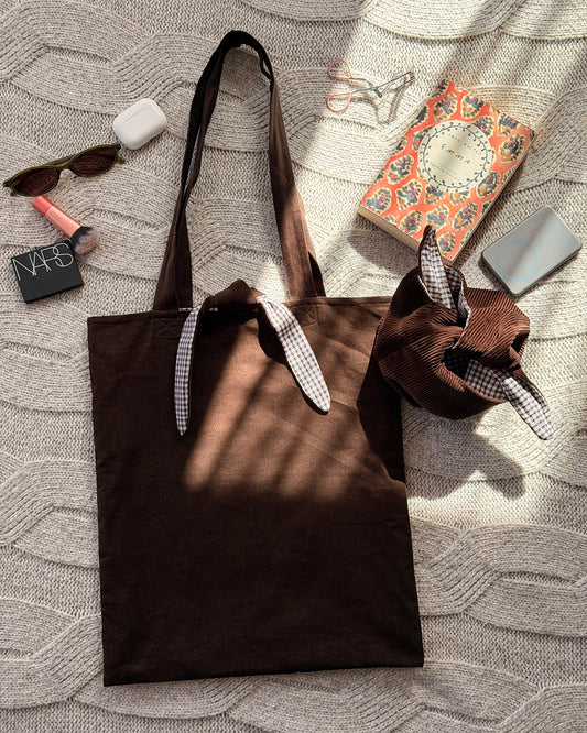 A dark chocolate brown corduroy fabric tote bag with long handles lined with brown gingham and tie and tie detail, with tie up organiser bag to match and beauty products scattered around. Laid flat on a textured cream knit blanket. Poetcore aesthetic.