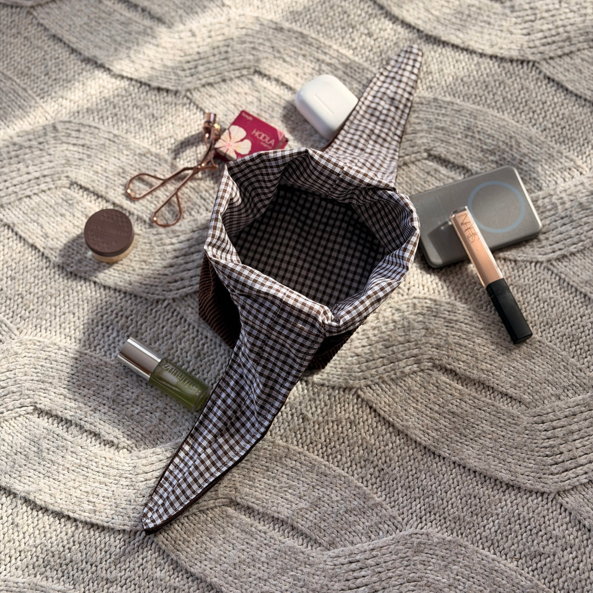 An empty styled flat-lay chocolate brown corduroy make up beauty organiser bag with a dark brown gingham lining with beauty products scattered around. Poetcore aesthetic. 