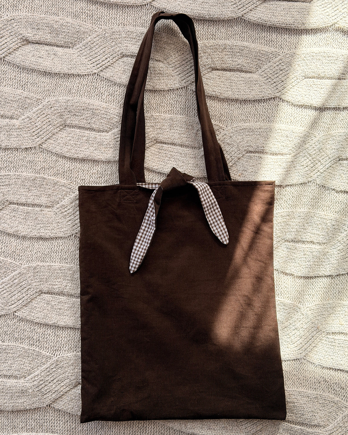 A dark chocolate brown corduroy fabric tote bag with long handles lined with brown gingham and tie and tie detail, laid flat on a textured cream knit blanket. Poetcore aesthetic.
