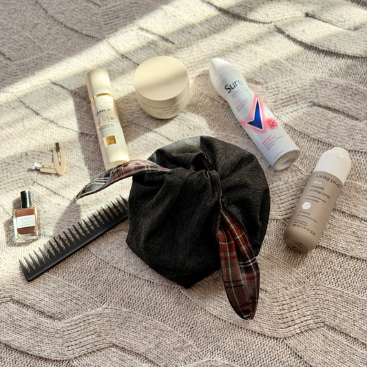 A styled flat-lay black denim make up beauty bag with a brown and grey tartan lining with beauty products scattered around. Dark Academia aesthetic. Surrounded by beauty products.