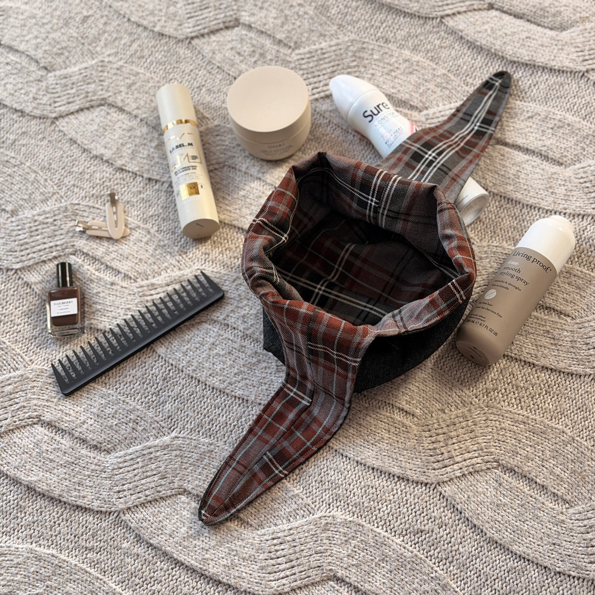 A empty styled flat-lay black denim make up beauty bag with a brown and grey tartan lining with beauty products scattered around. Dark Academia aesthetic. Surrounded by beauty products.