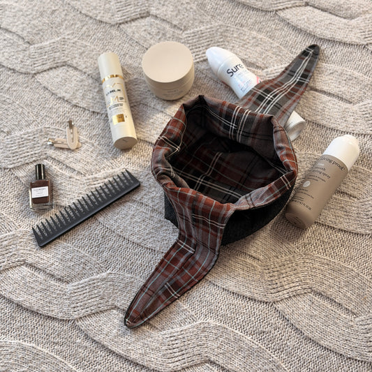 A empty styled flat-lay black denim make up beauty bag with a brown and grey tartan lining with beauty products scattered around. Dark Academia aesthetic. Surrounded by beauty products.