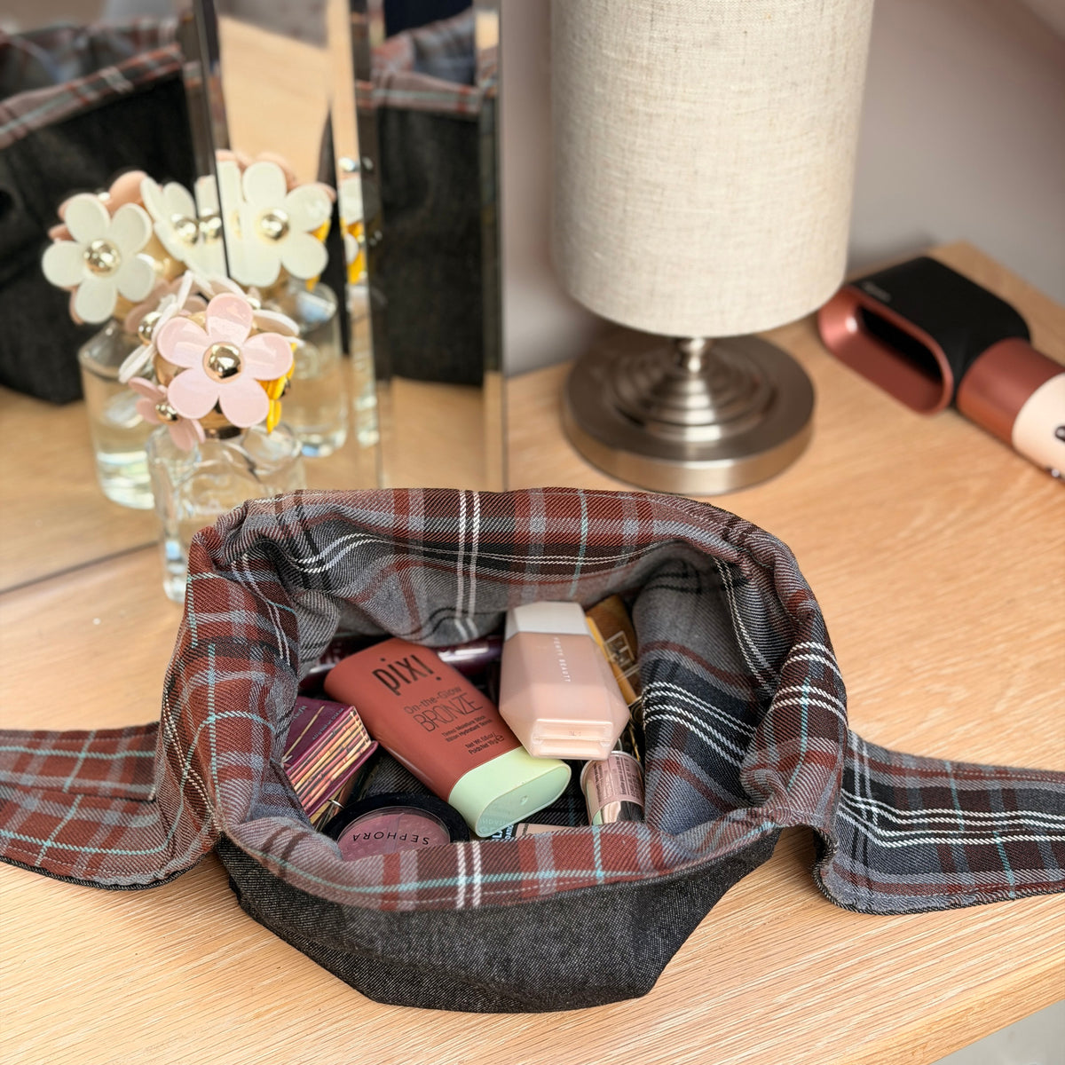 A fabric makeup bag with a brown and gray tartan pattern and a black denim base, filled with various cosmetic products, set on a wooden vanity table next to a lamp. Dark Academia aesthetic. 