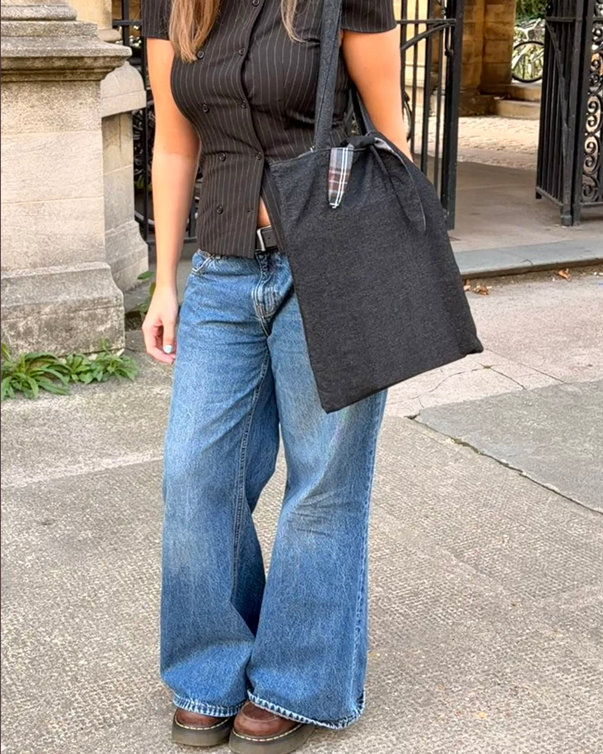 A person outdoors wearing a dark pinstriped short-sleeve top, wide-leg blue jeans, and brown shoes, carrying a large dark charcoal gray and tartan fabric tote bag over one shoulder. Dark Academia aesthetic.