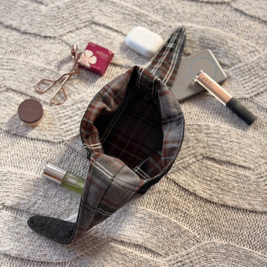 A styled flat-lay black denim empty make up organiser bag with a brown and grey tartan lining with beauty products scattered around. Dark Academia aesthetic. Surrounded by beauty products.
