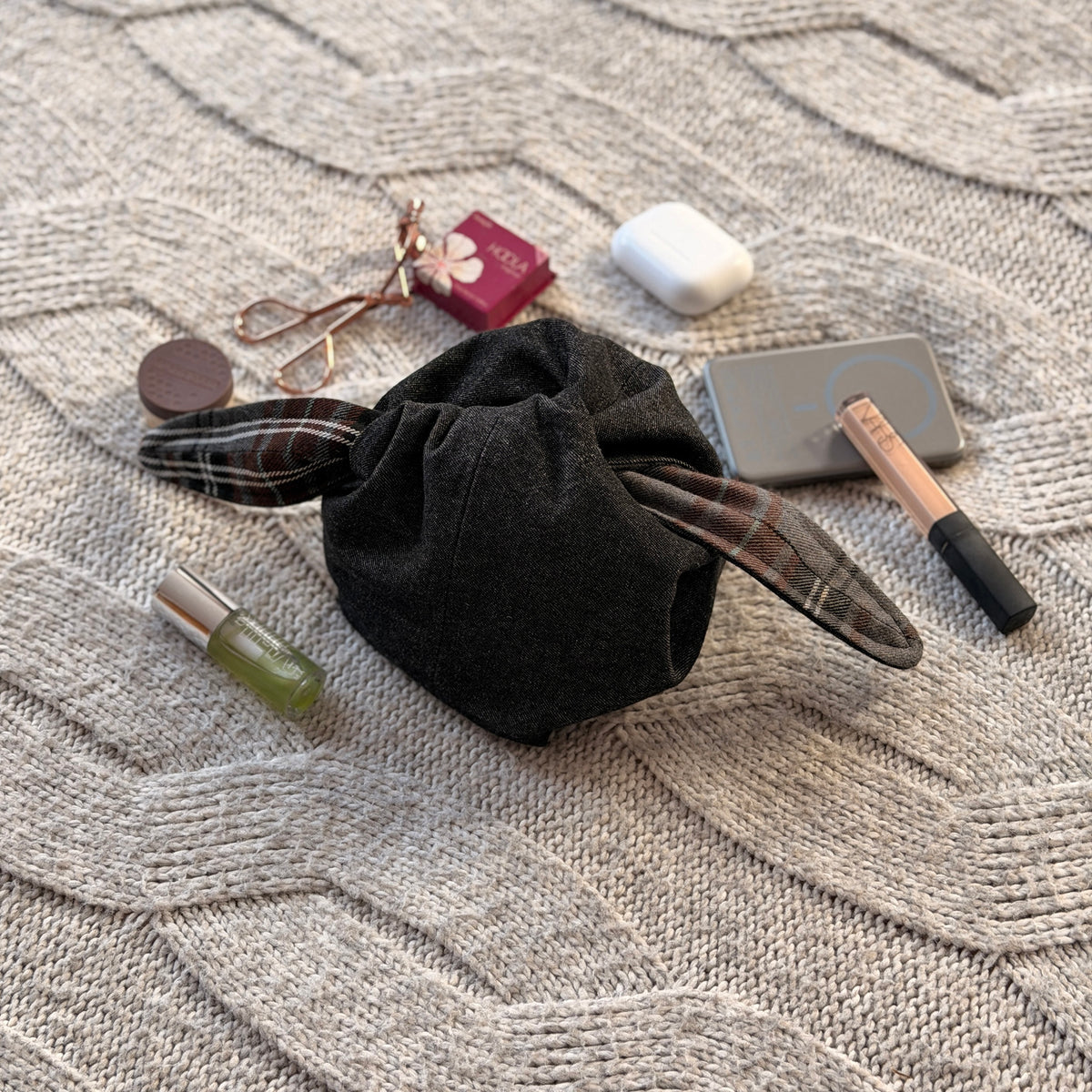 A styled flat-lay black denim make up organiser bag with a brown and grey tartan lining with beauty products scattered around. Dark Academia aesthetic. Surrounded by beauty products. 