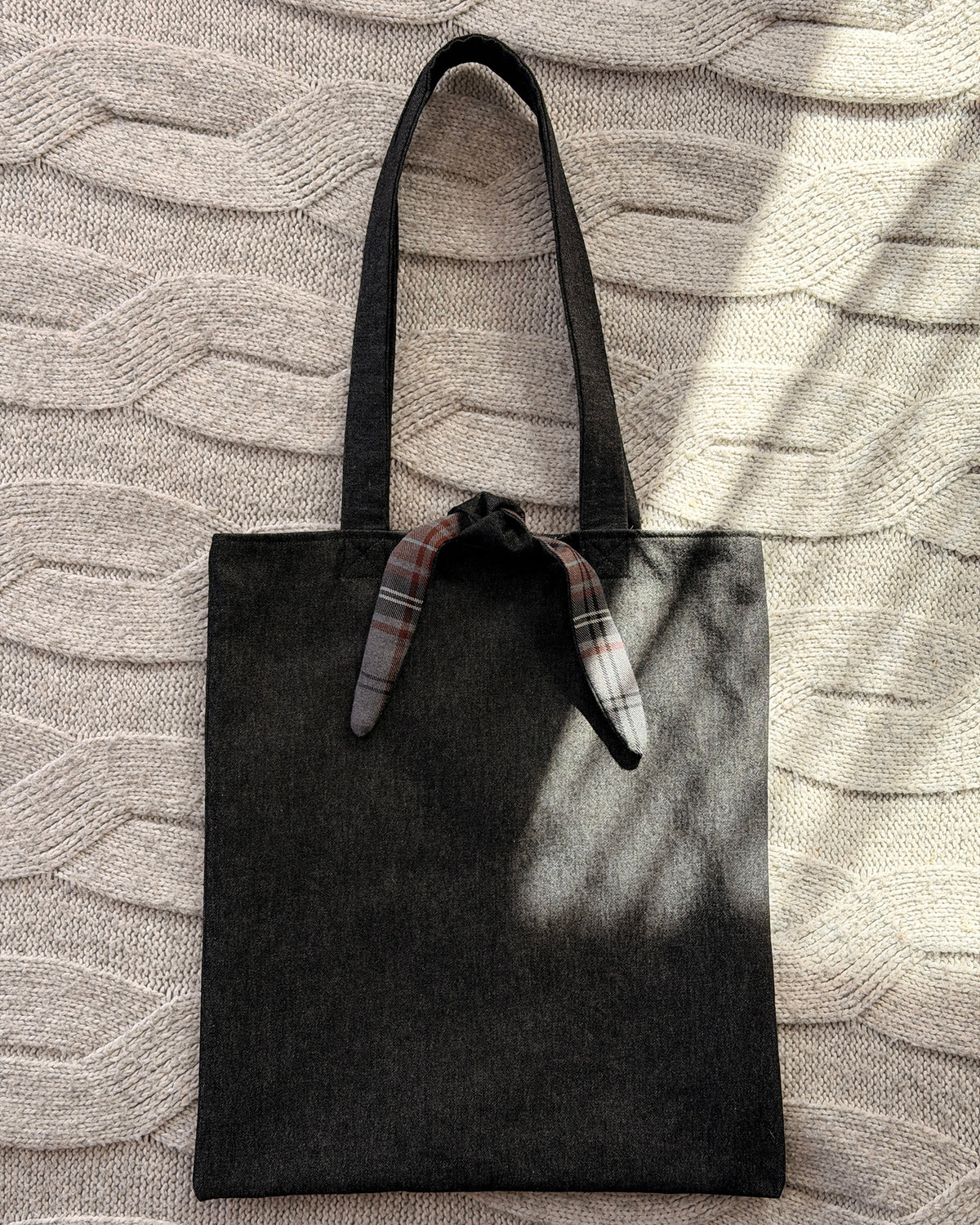 A dark charcoal denim fabric tote bag with black shoulder straps and a tartan tie detail at the top, laid flat on a textured beige knit blanket with dappled sunlight. Dark Academia aesthetic.