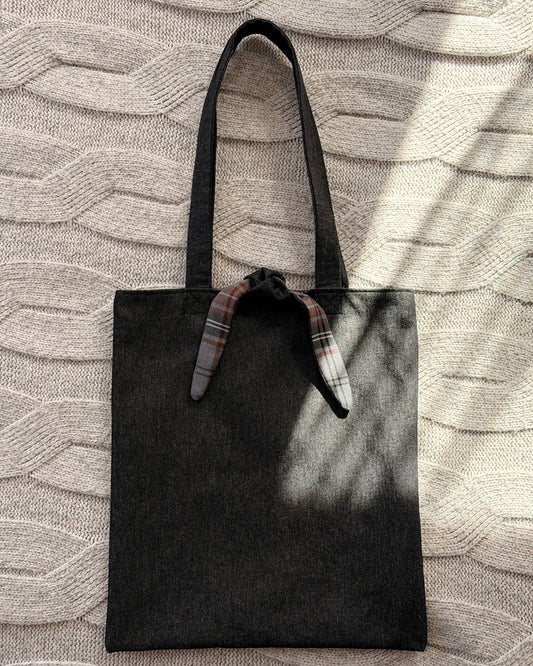 A dark charcoal denim fabric tote bag with black shoulder straps and a tartan tie detail at the top, laid flat on a textured beige knit blanket with dappled sunlight. Dark Academia aesthetic.