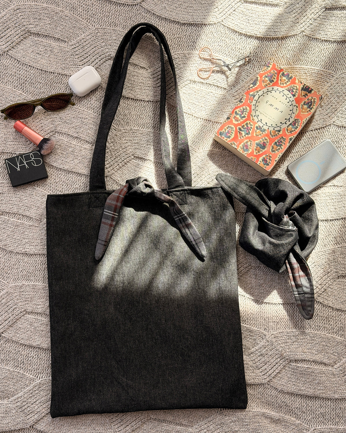 A dark charcoal denim fabric tote bag with black shoulder straps and a tartan tie detail at the top, laid flat on a textured beige knit blanket with dappled sunlight. Dark Academia aesthetic. With lifestyle fabrics scattered around.