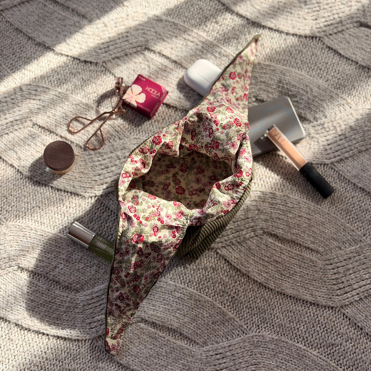 An empty styled flat-lay olive green corduroy make up organiser bag with a pink ditsy floral lining with beauty products scattered around. Cottagecore aesthetic. Surrounded by beauty products