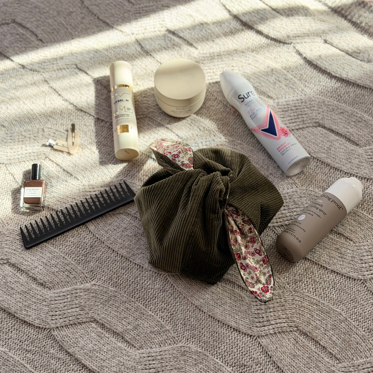A styled flat-lay olive green corduroy make up beauty bag with a pink ditsy floral lining with beauty products scattered around. Cottagecore aesthetic. Surrounded by beauty products. 