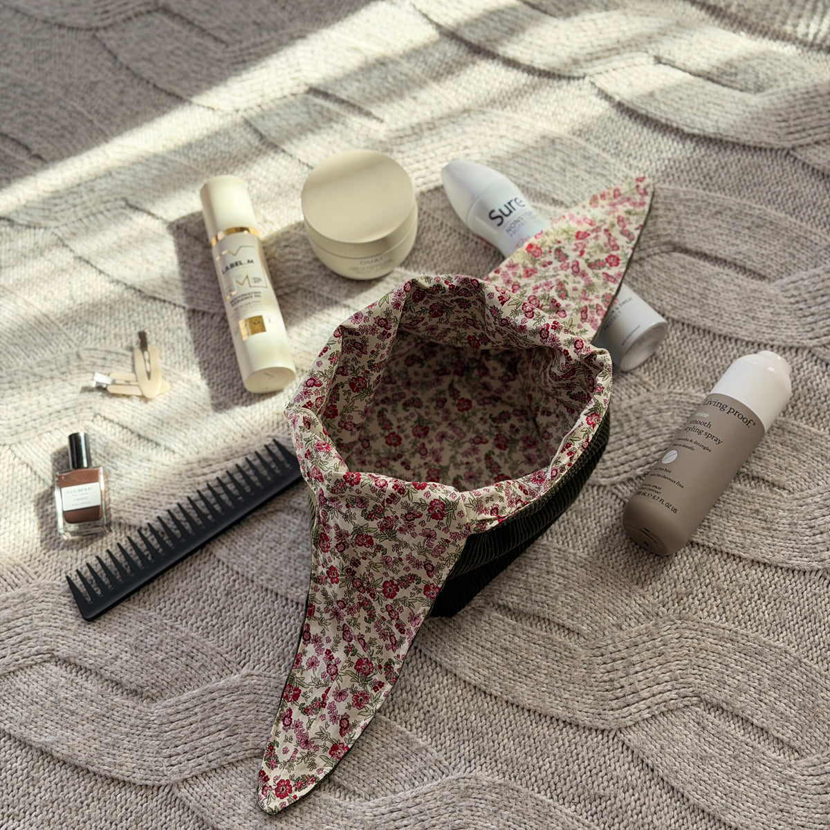 A styled flat-lay empty olive green corduroy make up beauty bag with a pink ditsy floral lining with beauty products scattered around. Cottagecore aesthetic. Surrounded by beauty products.