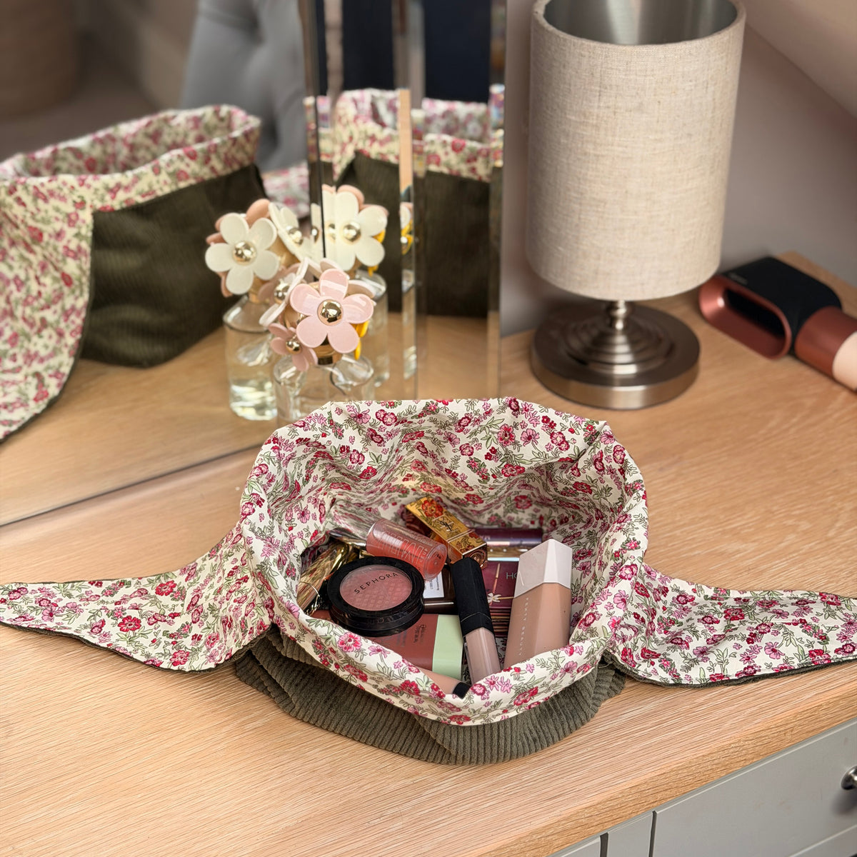 A flat lay olive green corduroy fabric cosmetic bag with a pink and green ditsy floral print and olive green trim, filled with makeup items on a wooden vanity table next to a mirror and lamp. Cottagecore aesthetic.