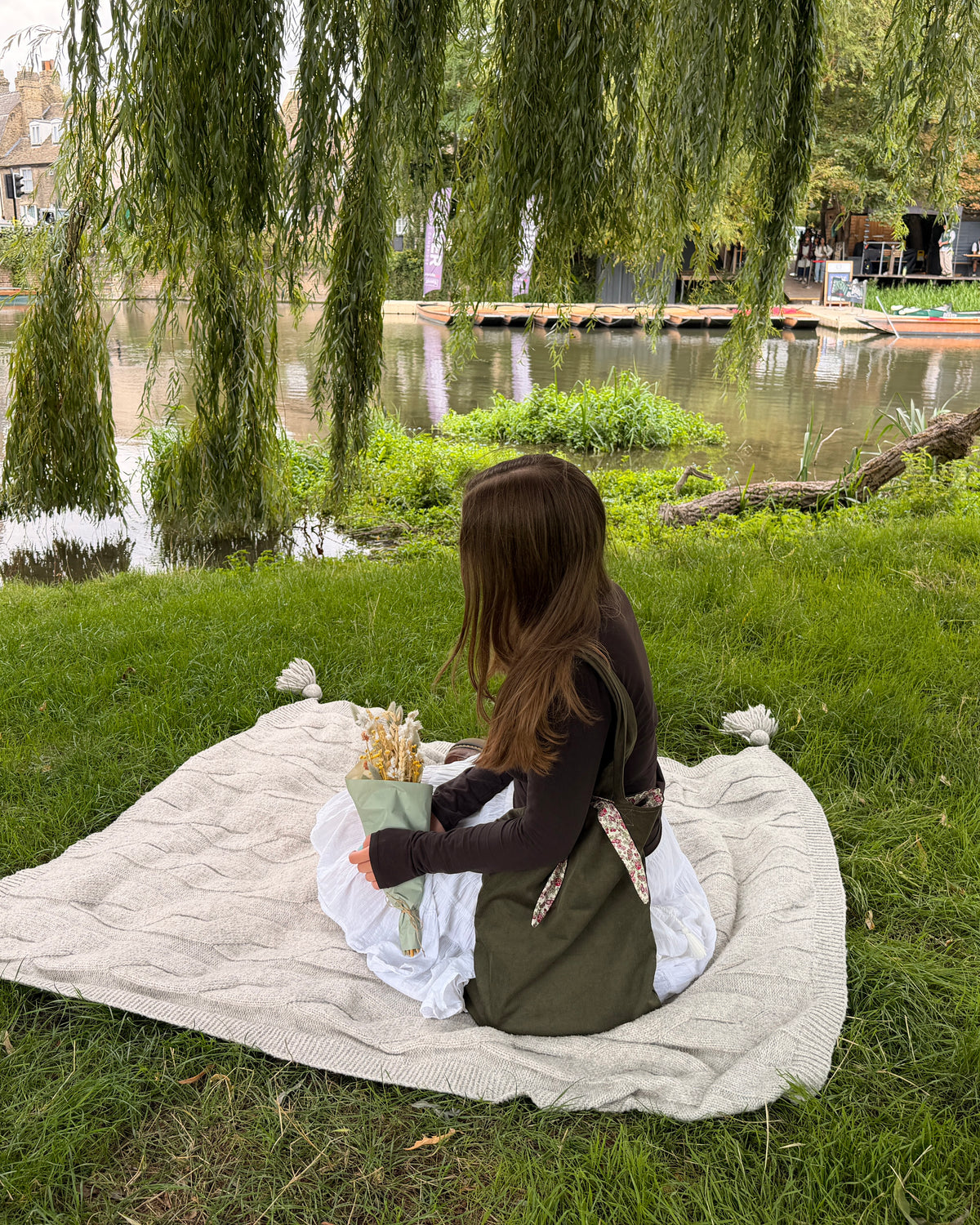 A woman sits on a cream knit blanket on a grassy lawn by The Granta river Cambridge, holding a bouquet of dried flowers. She is wearing a dark brown long-sleeve top, an olive green apron, and a white skirt. Cottagecore aesthetic