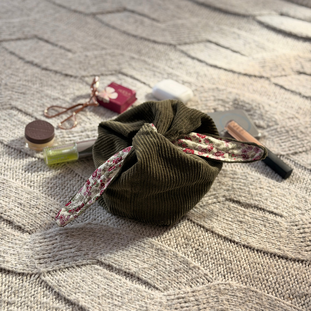 
A styled flat-lay olive green corduroy make up organiser bag with a pink ditsy floral lining with beauty products scattered around. Cottagecore aesthetic. Surrounded by beauty products