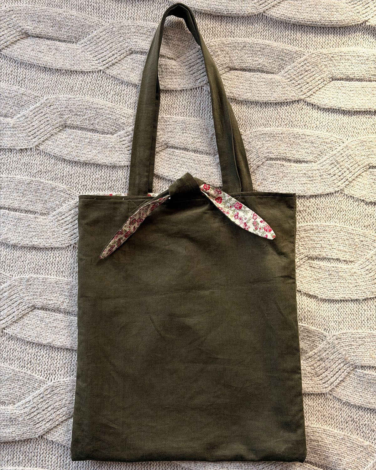 An olive green canvas tote bag with pink ditsy floral floral-patterned fabric accents on the handles and top trim, laid flat on a textured gray knit blanket. Cottagecore aesthetic.