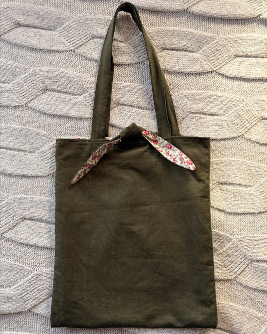 An olive green canvas tote bag with pink ditsy floral floral-patterned fabric accents on the handles and top trim, laid flat on a textured gray knit blanket. Cottagecore aesthetic.
