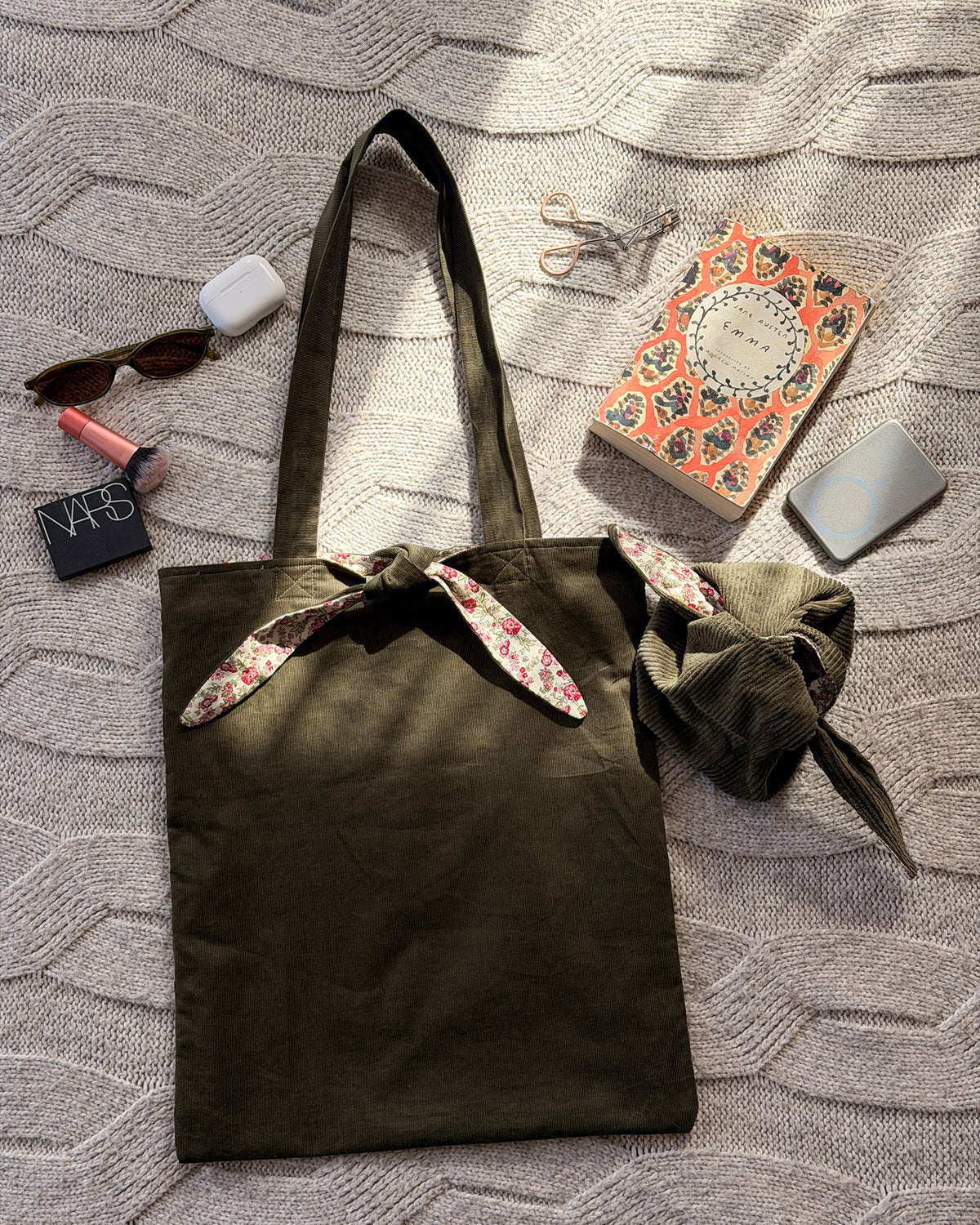 An olive green canvas tote bag with pink ditsy floral floral-patterned fabric accents on the handles and top trim, laid flat on a textured gray knit blanket. Cottagecore aesthetic. With lifestyle products scattered around.