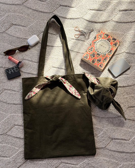 An olive green canvas tote bag with pink ditsy floral floral-patterned fabric accents on the handles and top trim, laid flat on a textured gray knit blanket. Cottagecore aesthetic. With lifestyle products scattered around.