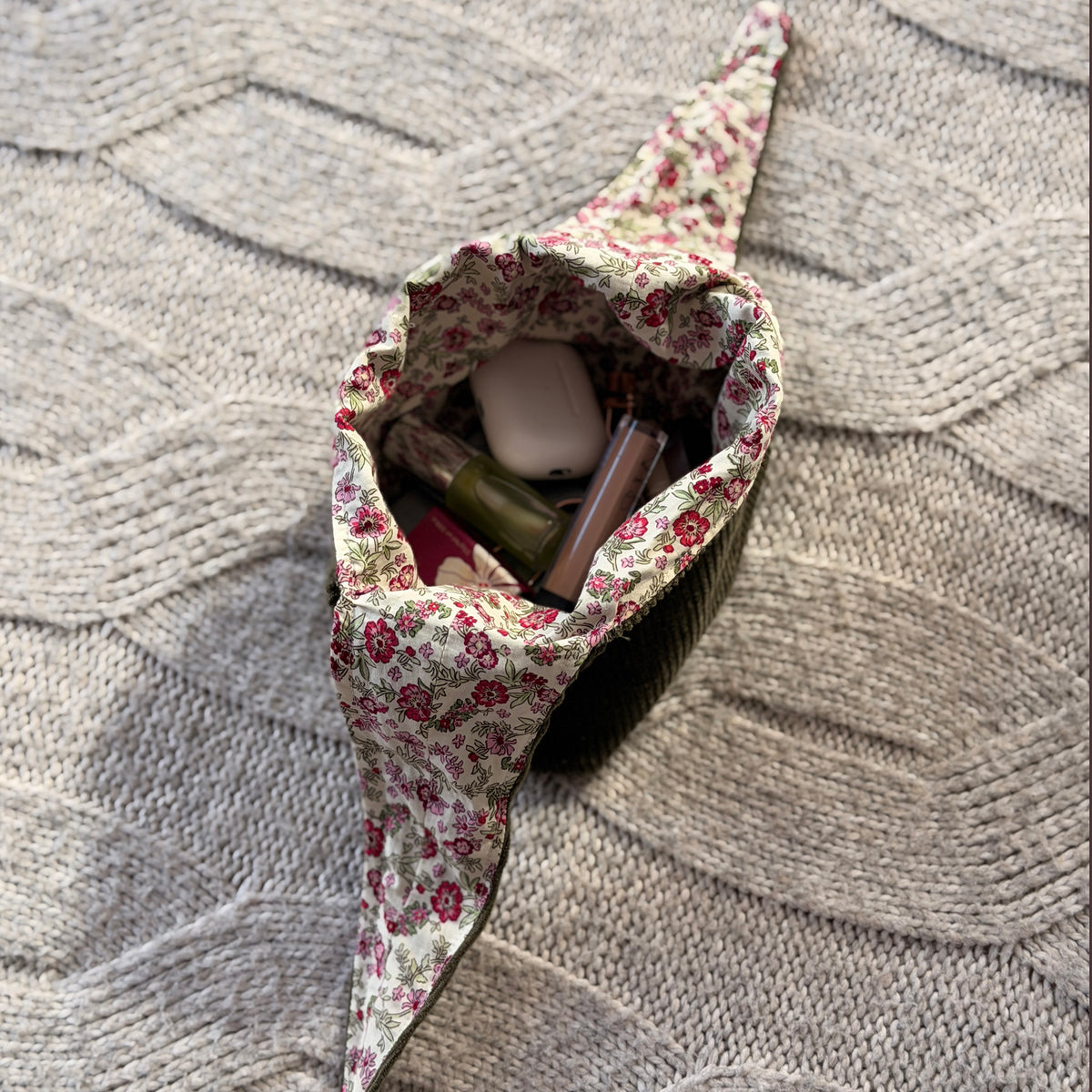 A filled styled flat-lay olive green corduroy make up organiser bag with a pink ditsy floral lining with beauty products scattered around. Cottagecore aesthetic. Surrounded by beauty products