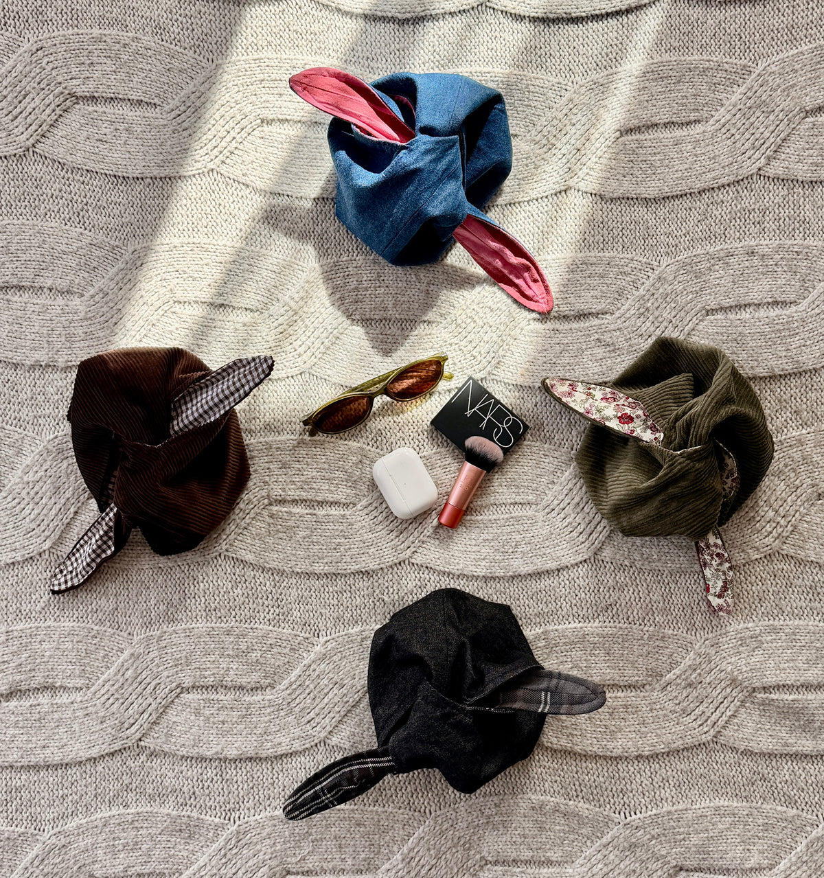 A styled flat-lay of four tie up make up organiser bags. dark brown corduroy, washed black denim, denim blue, olive green corduroy, and pink. Alongside Airpods, sunglasses and make up
