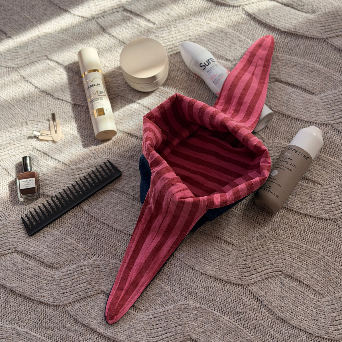 A styled flat-lay dark denim make up beauty bag with striped pink and maroon lining with beauty products scattered around. Y2k aesthetic.