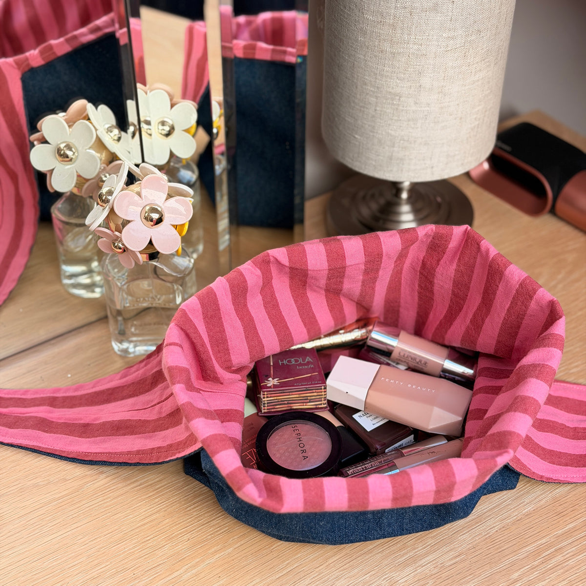 A pink and maroon striped fabric flay lay makeup bag filled with cosmetics, placed on a wooden desk next to a lamp and decorative flowers. Y2K aesthetic.