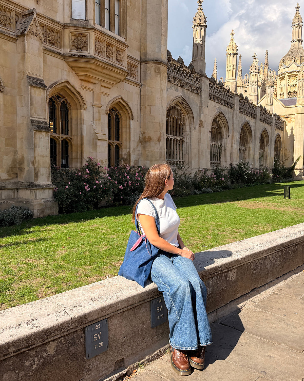 A woman sits on a stone ledge outdoors in front of a Kings College Cambridge and green grass behind her. She is wearing a white top, wide-leg blue jeans, brown shoes, and has a dark denim tie up tote bag. Y2K aesthetic.